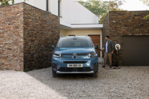 A blue Citroën van stands in front of a modern house while a man holds a charging cable in his hand.