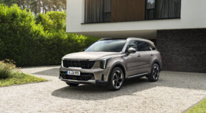 A Kia Sorento stands on a bright gravel driveway in front of a modern building with large windows and a green hedge in the background.