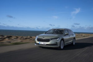 The picture shows a silver Škoda Superb Combi driving on a coastal road.