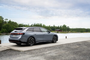 Silver VW ID.7 Tourer, photographed from diagonally behind, parked on a lakeshore with a forest and a wooden house in the background.