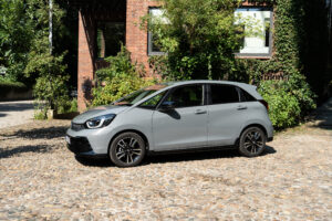 A gray Honda Jazz is parked in a cobbled square in front of a green brick building.
